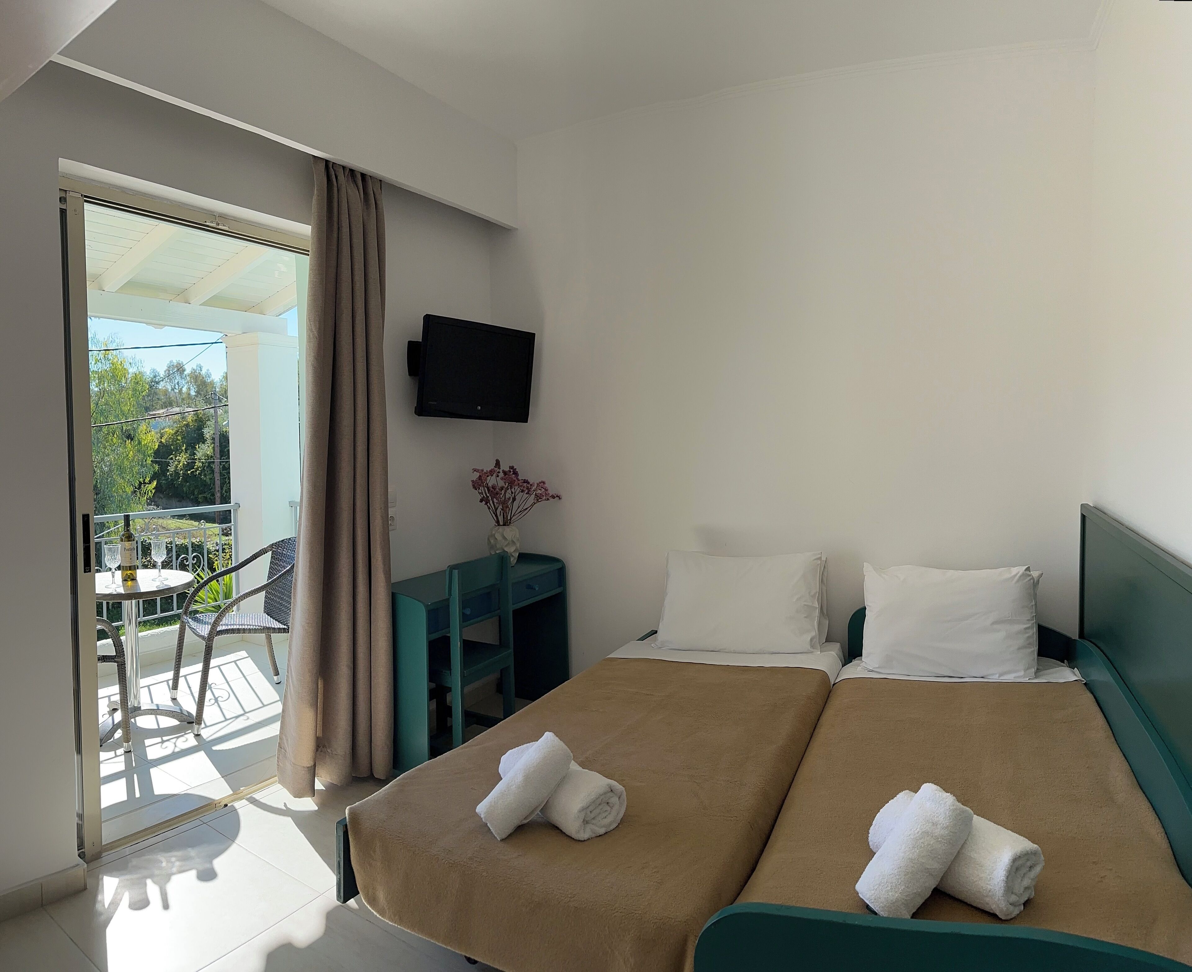 A hotel room with two beds, a TV, a small table with a bottle, and a view of greenery outside.