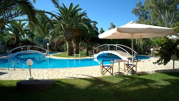 Relax by the serene pool surrounded by lush greenery and palm trees, with comfortable seating under a large umbrella.