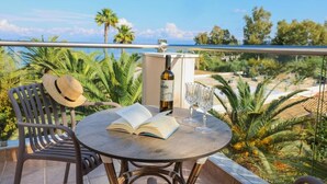 Una mesa redonda con un libro abierto y dos copas de vino, un sombrero de paja y una botella de vino en un balcón con vistas a palmeras y una playa.