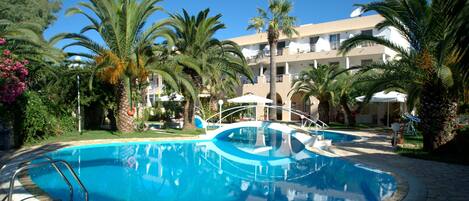 A circular swimming pool with a clear blue water surface, surrounded by palm trees and a building with balconies in the background.