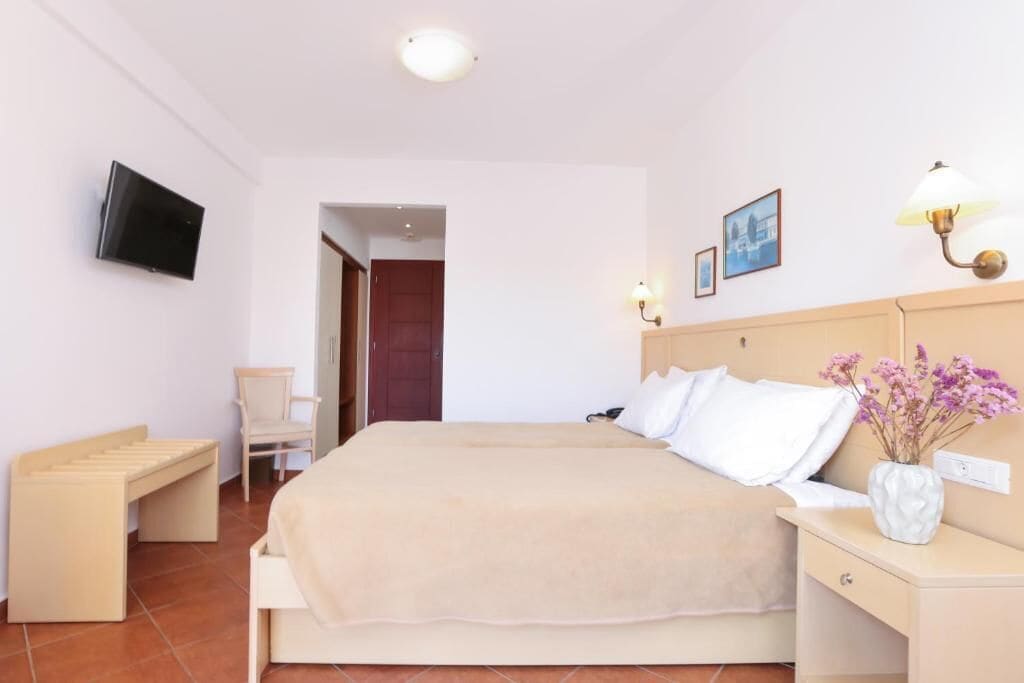 Una habitación de hotel con una cama, una silla, una mesita de noche con lámpara, un televisor montado en la pared y un cuadro en la pared.