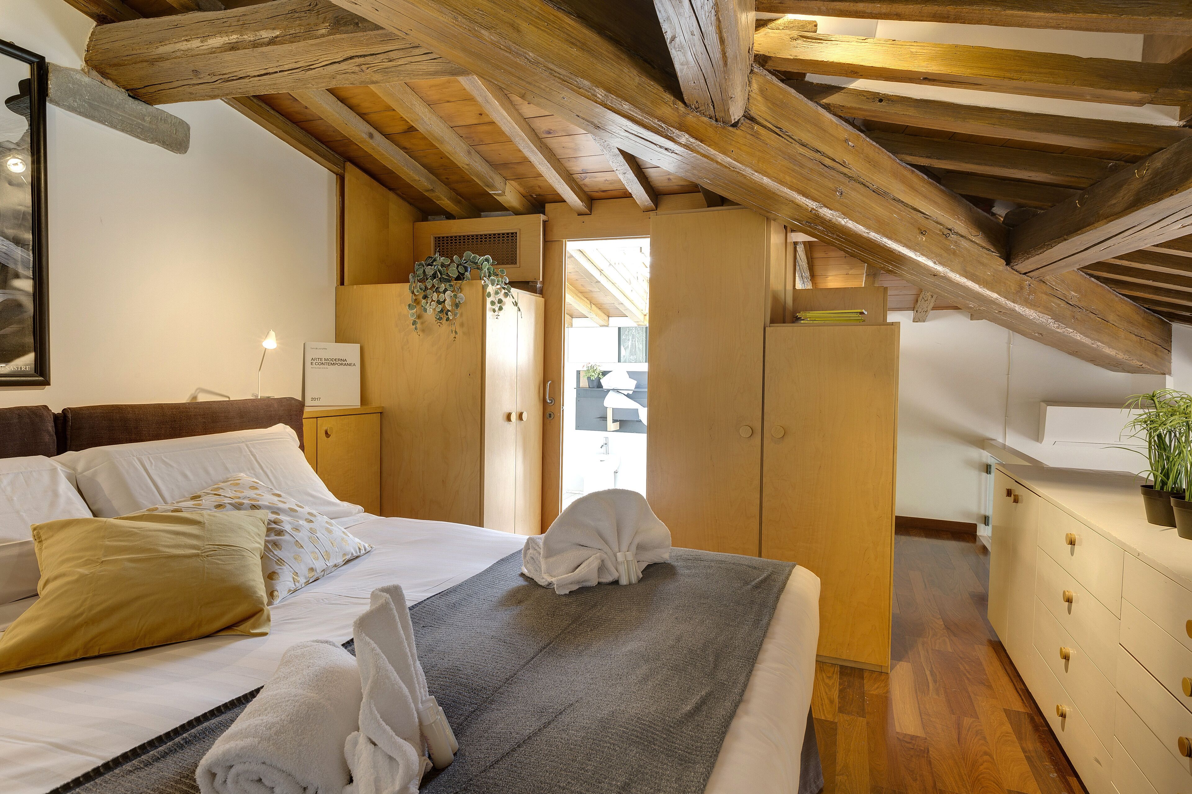 A hotel room with a wooden ceiling, a bed with white linens, a wooden wardrobe, and a small plant on the dresser.