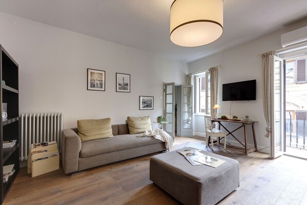 Un salon moderne avec un canapé, un pouf et une table sur laquelle se trouve une lampe. Un téléviseur est fixé au mur et un radiateur se trouve à gauche.