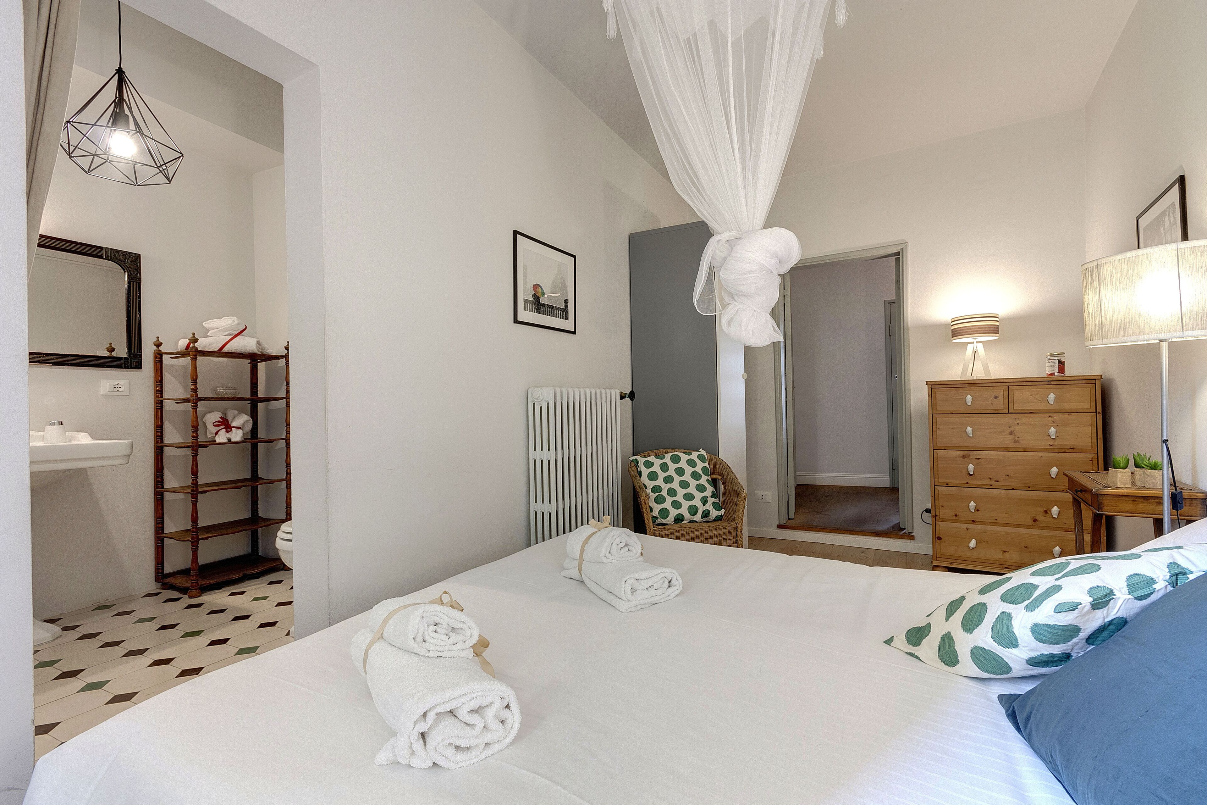 A bedroom with a bed, a wooden dresser, a chair, a mirror, and a bathroom with a toilet and shelves.