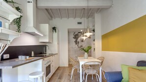 A compact kitchen with white cabinetry, a dining area with wicker chairs, and a sofa with cushions. - Mamo Florence - Ricasoli Apartment (Florence)