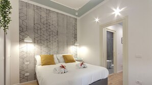 A neatly made bed with white linens and two rolled towels, a geometric-patterned wall, and a hanging pendant light. - Mamo Florence - Ricasoli Apartment (Florence)