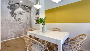 A dining area with a white table, wicker chairs, a white vase with a plant, a mural on the wall, and a yellow accent wall. - Mamo Florence - Ricasoli Apartment (Florence)