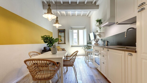 A modern kitchen with white cabinetry, a dining area with wicker chairs, and a large window providing natural light. - Mamo Florence - Ricasoli Apartment (Florence)