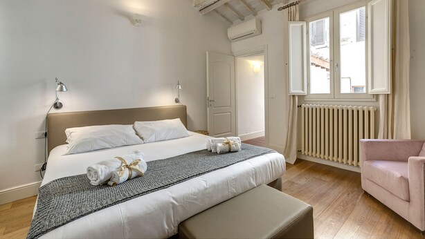 A bedroom with a large bed, bedside lamps, a radiator, a window with curtains, and a pink chair. - Mamo Florence - Tegolaio Apartment (Florence)