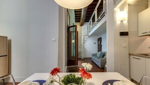 A dining area with a table set for two, a vase with flowers, and a view into a living room with a sofa and a staircase. - Mamo Florence - Tegolaio Apartment (Florence)