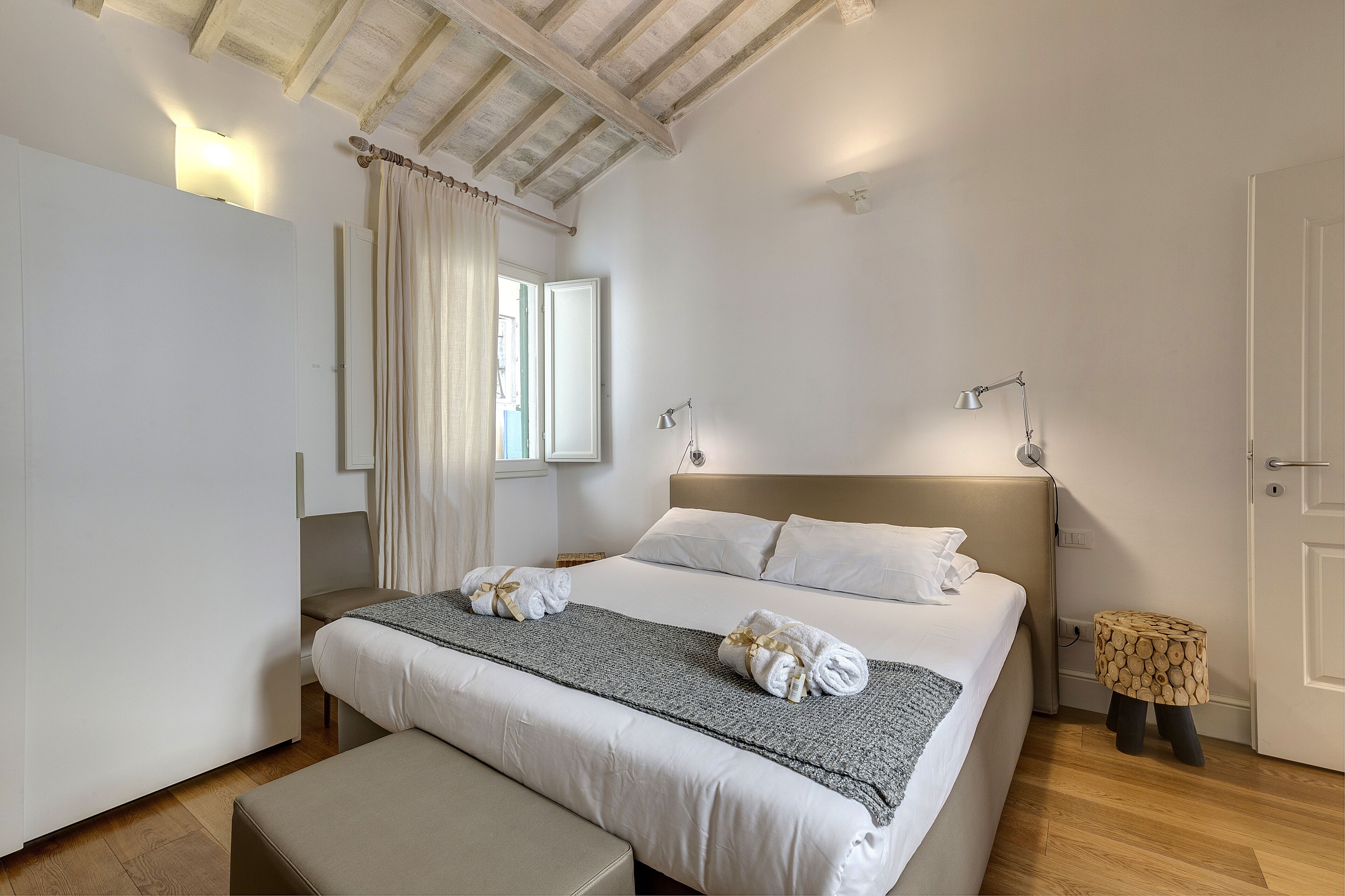 A bedroom with a large bed, a bedside table, a bench, and a window with curtains.