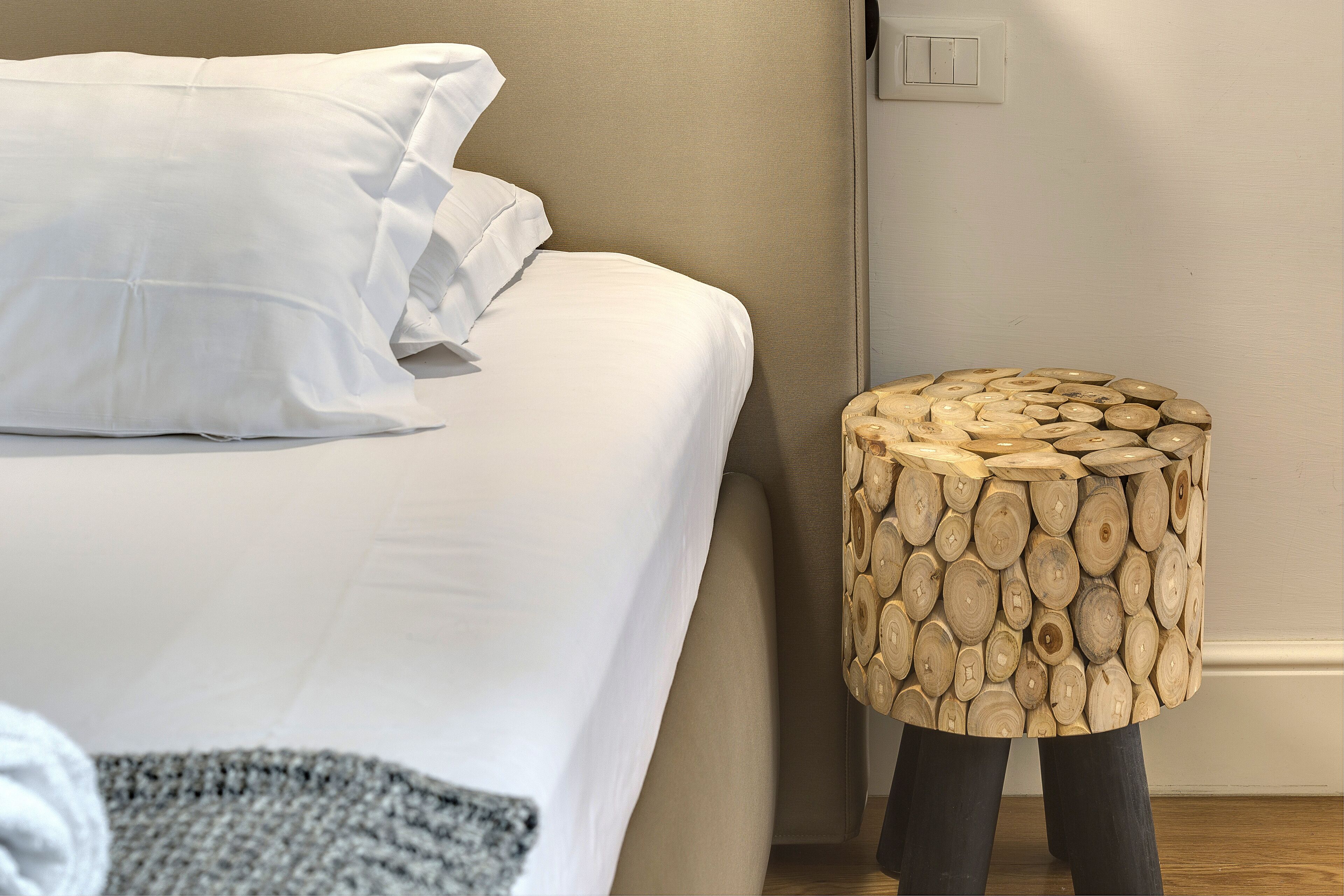 Un lit bien fait, avec du linge de lit blanc, et un tabouret rond en bois à la finition naturelle.