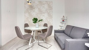 Una sala moderna con un sofá gris, una mesa redonda de cristal y cuatro sillas grises, una planta en maceta y un estante en la pared con libros.