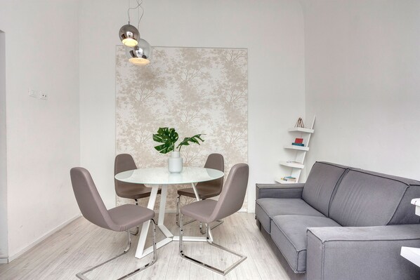 Una sala moderna con un sofá gris, una mesa redonda de cristal y cuatro sillas grises, una planta en maceta y un estante en la pared con libros.