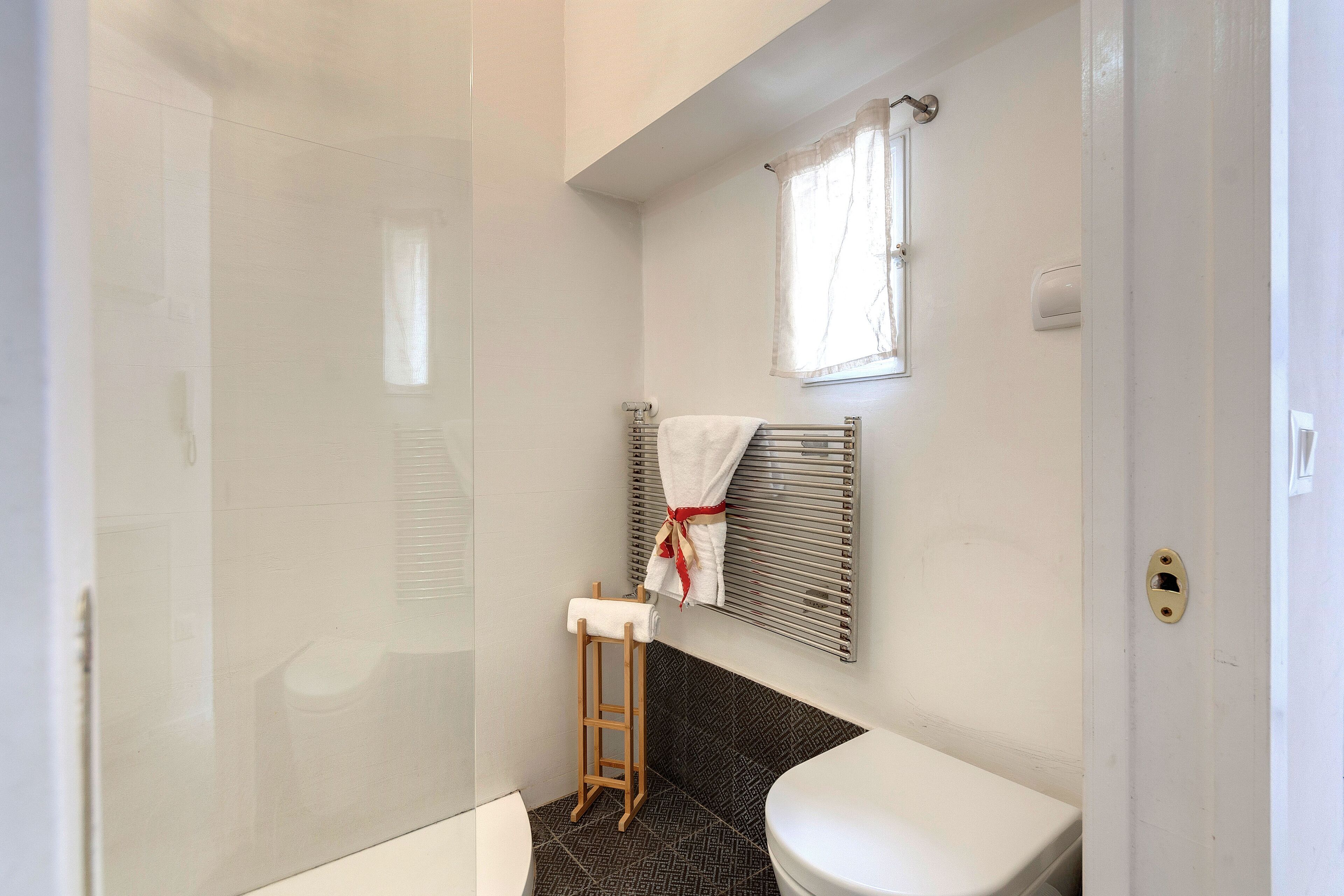 Baño con cabina de ducha de vidrio, inodoro blanco, un taburete pequeño de madera, toallero con toalla y ventana con persianas.