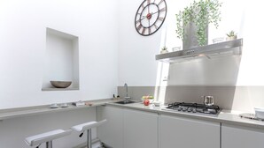 Una cocina moderna con estufa a gas incorporada, un lavamanos y un reloj en la pared. Hay dos taburetes de bar y una pequeña estantería con un cuenco y una planta.