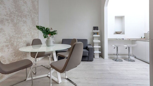 Un comedor moderno con una mesa de vidrio, dos sillas, una planta en maceta, un sofá gris y una zona de cocina con un estante y un cuenco.