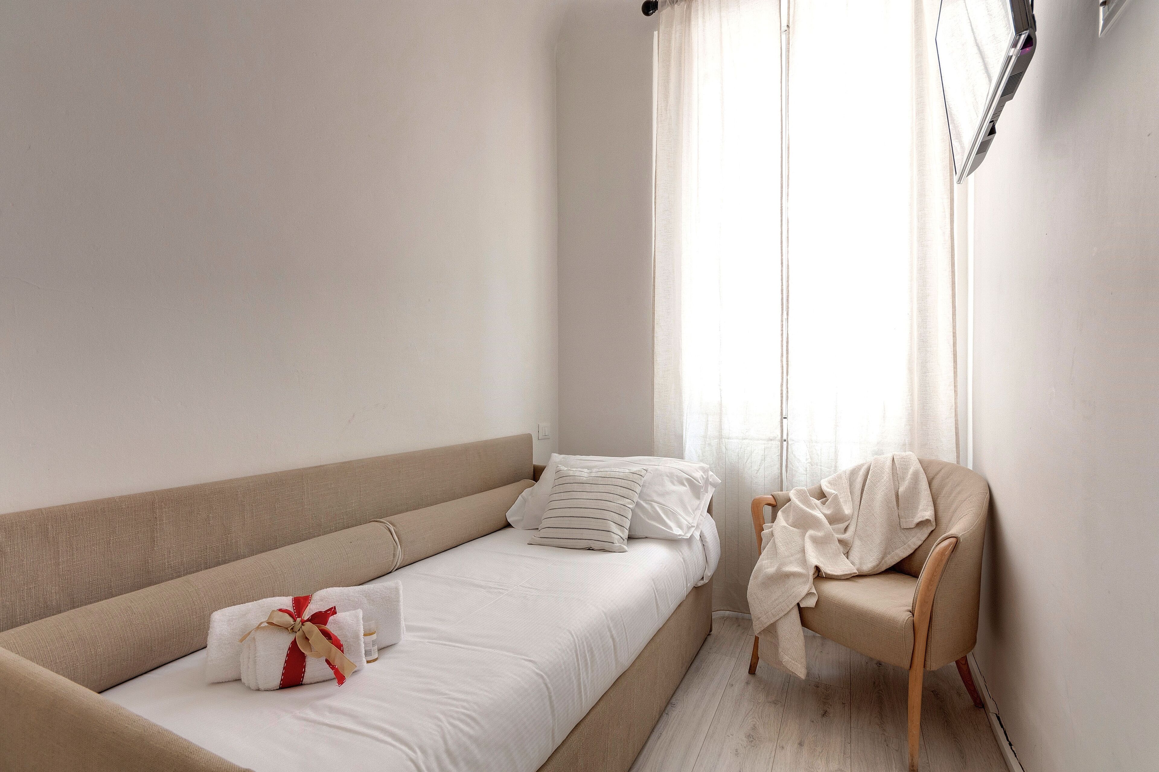 A minimalist hotel room with a single bed, a chair, and a window with sheer curtains.