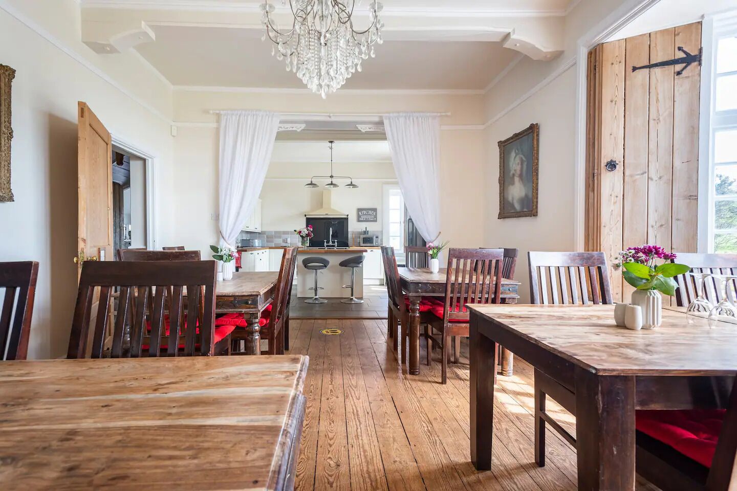 Un espace repas avec des tables et des chaises en bois, un lustre et une vue sur la cuisine.