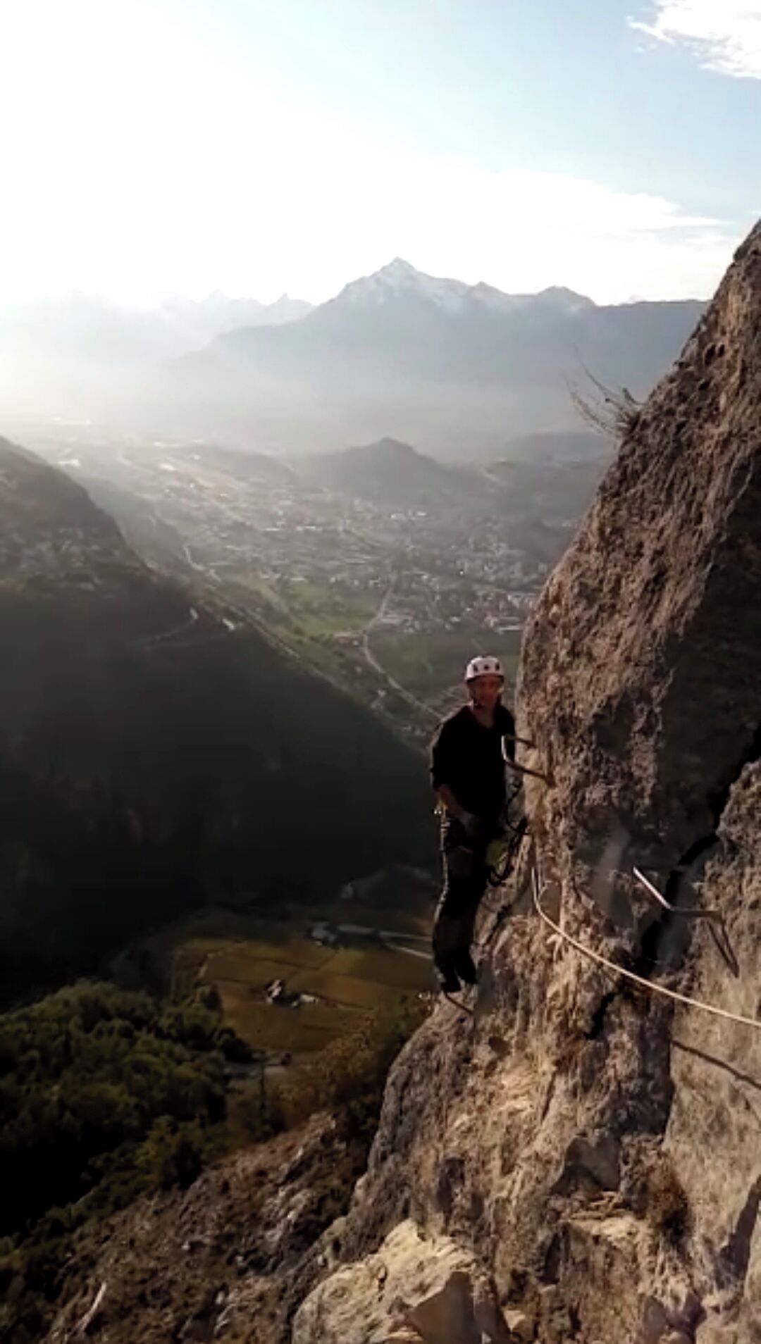 A person rappelling down a steep mountain cliff with a rope, overlooking a valley with buildings and mountains in the background.