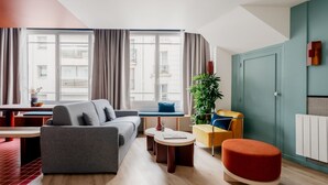 A modern living room with a grey sofa, a red ottoman, a yellow chair, and a blue bench. There is a large window with curtains, a plant, and a radiator on the wall.