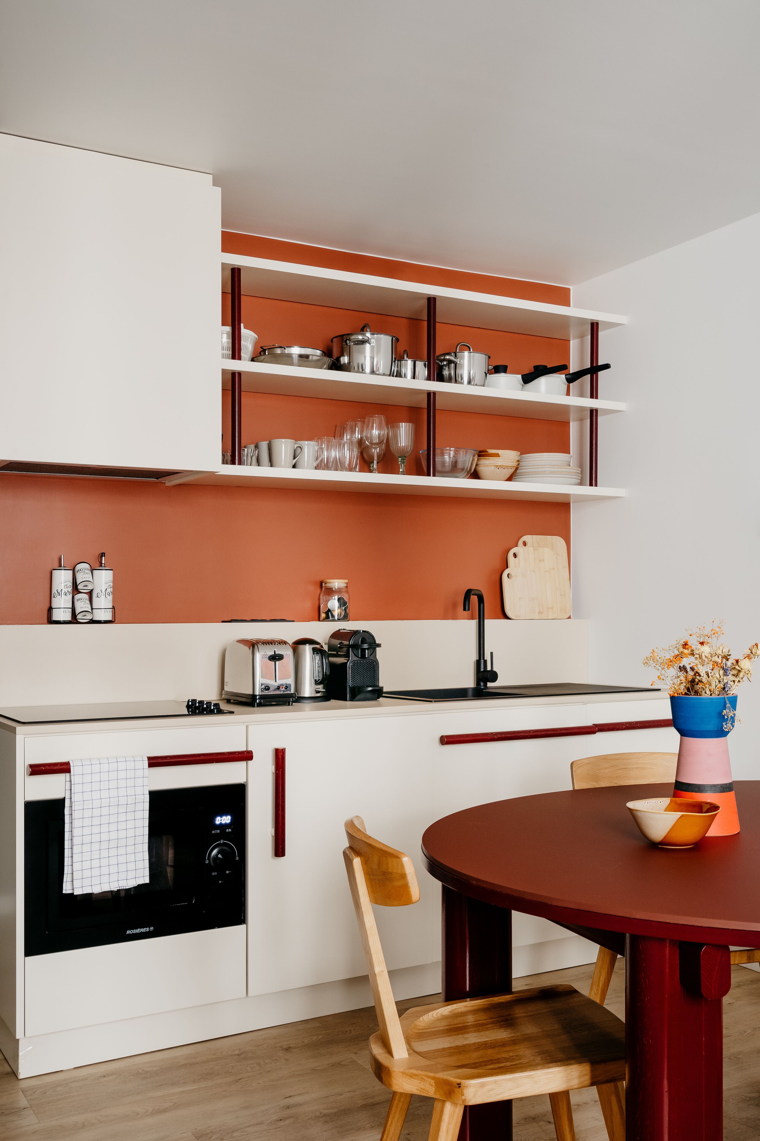 A compact kitchen with white cabinets, a built-in oven, a sink, and a round dining table with a chair.