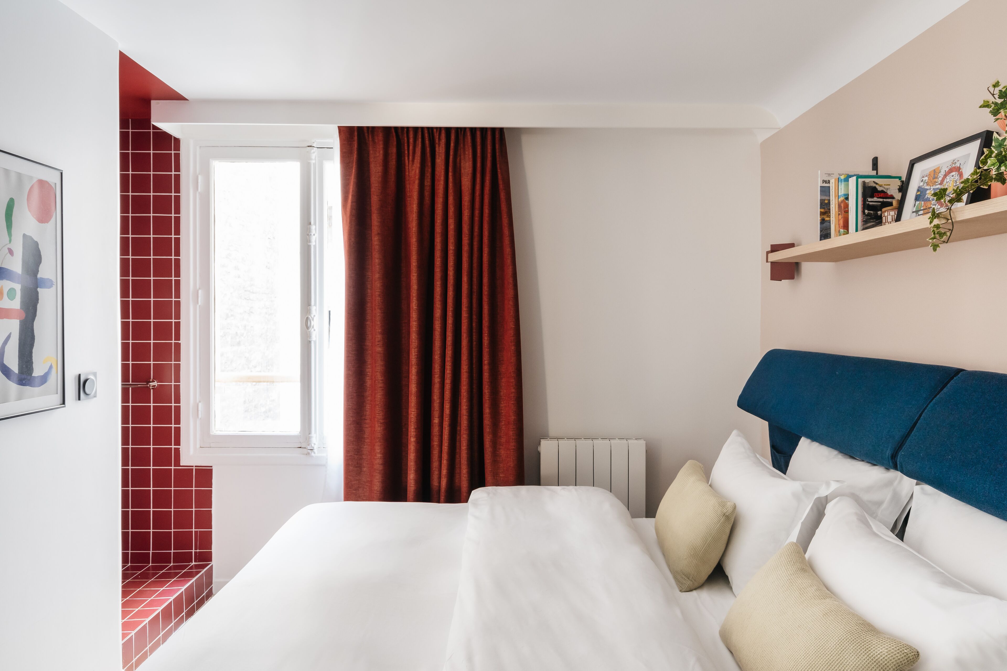 A bedroom with a bed, red curtains, a radiator, a shelf with books and a plant, and a red-tiled wall.