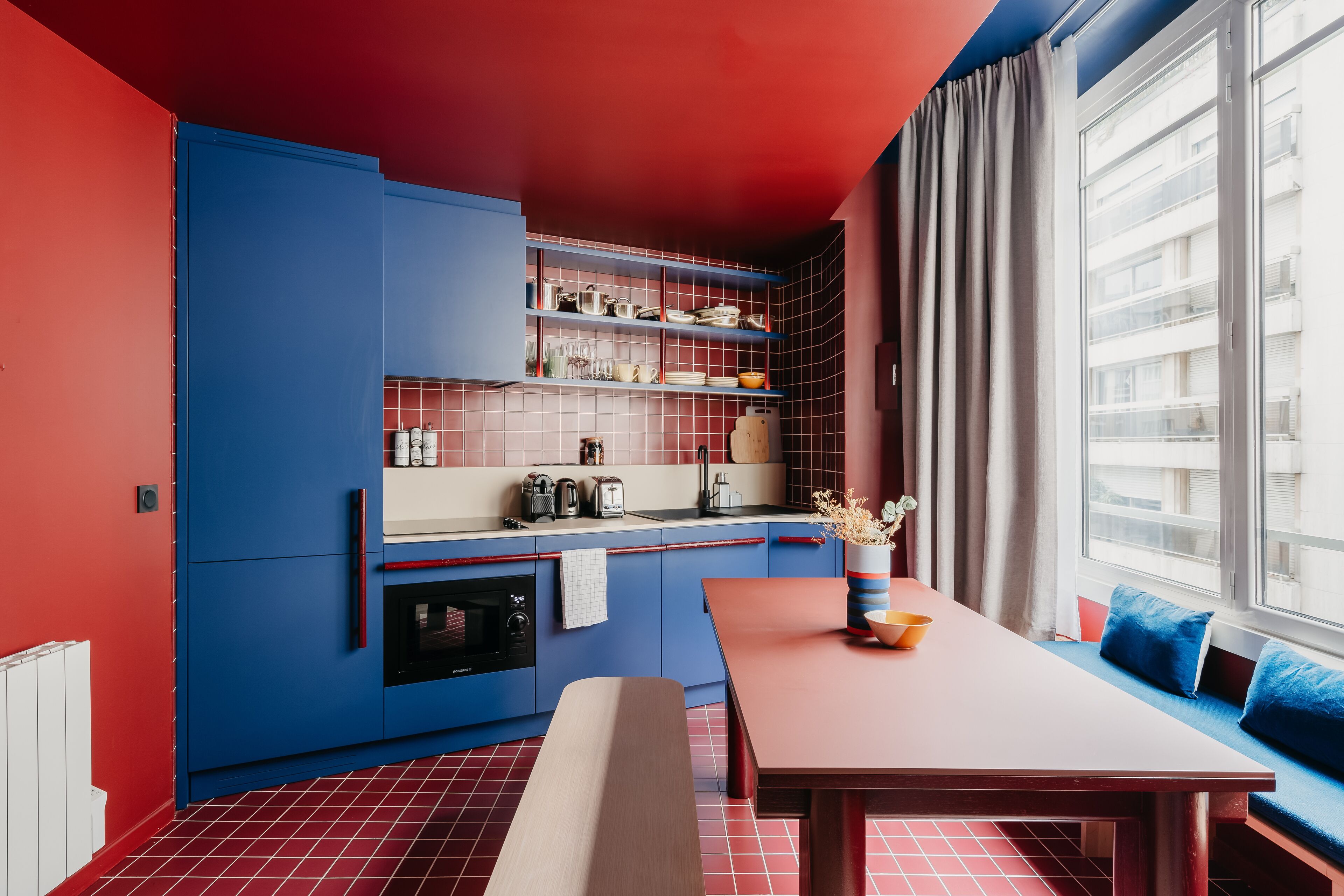 Una cocina moderna con armarios azules, techo rojo y una ventana con vistas a edificios.