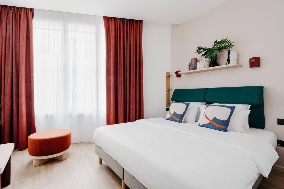 Una habitación de hotel moderna con una cama grande, un reposapiés rojo, una mesita de noche de madera y un estante montado en la pared con objetos decorativos.