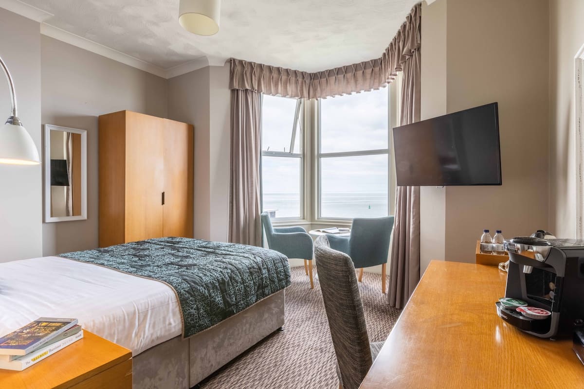 A hotel room with a bed, a desk with a coffee maker, a TV, and a view of the sea through large windows.