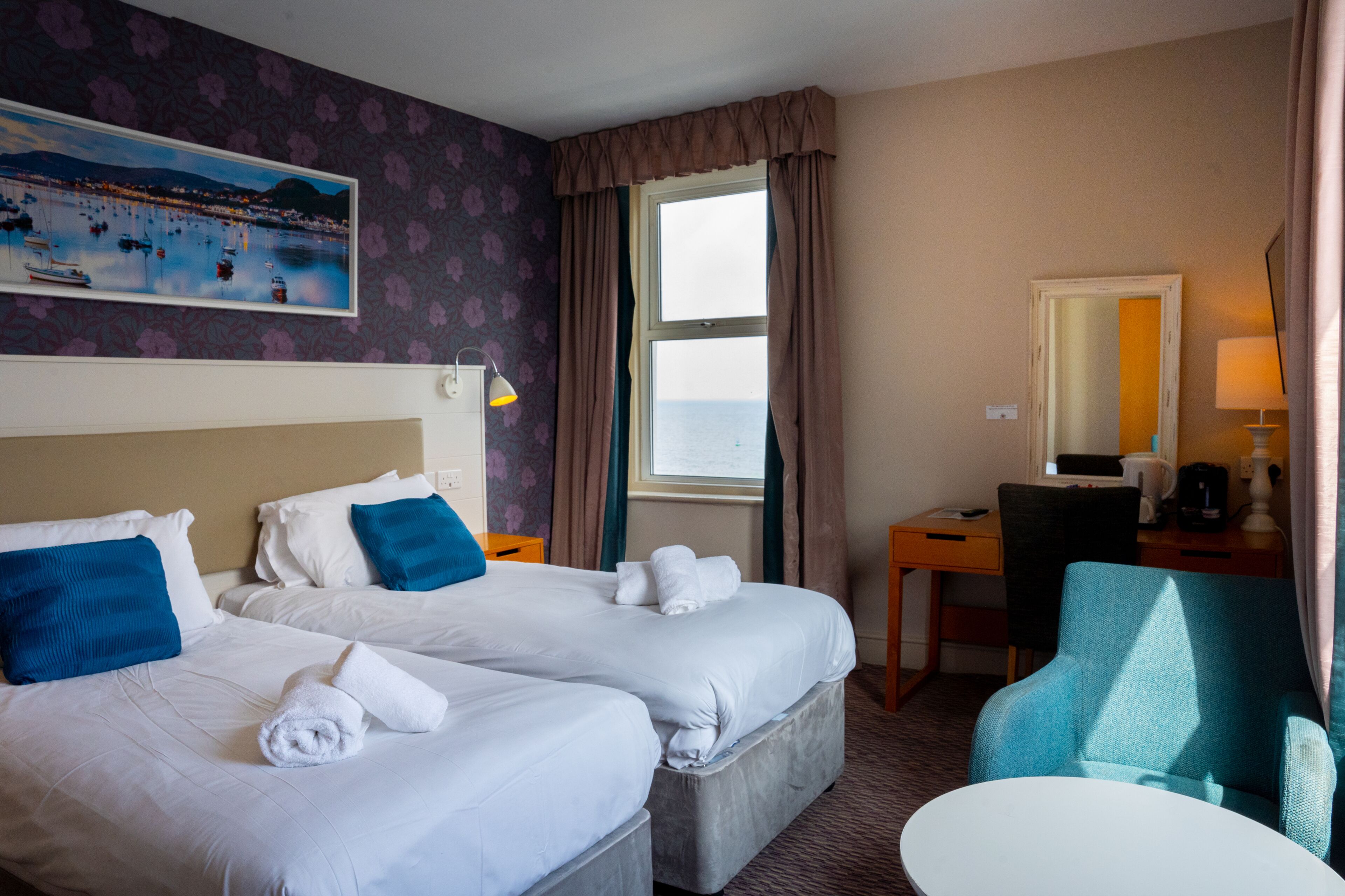 A hotel room with two beds, a desk, a chair, a window with curtains, and a painting on the wall.