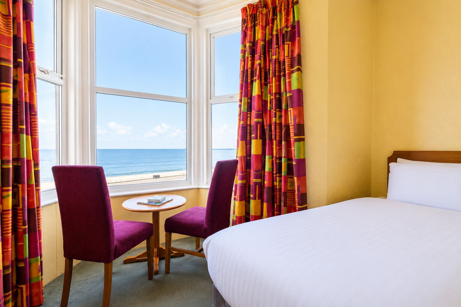 A hotel room with a bed, two chairs, a small table, and a view of the sea through large windows.