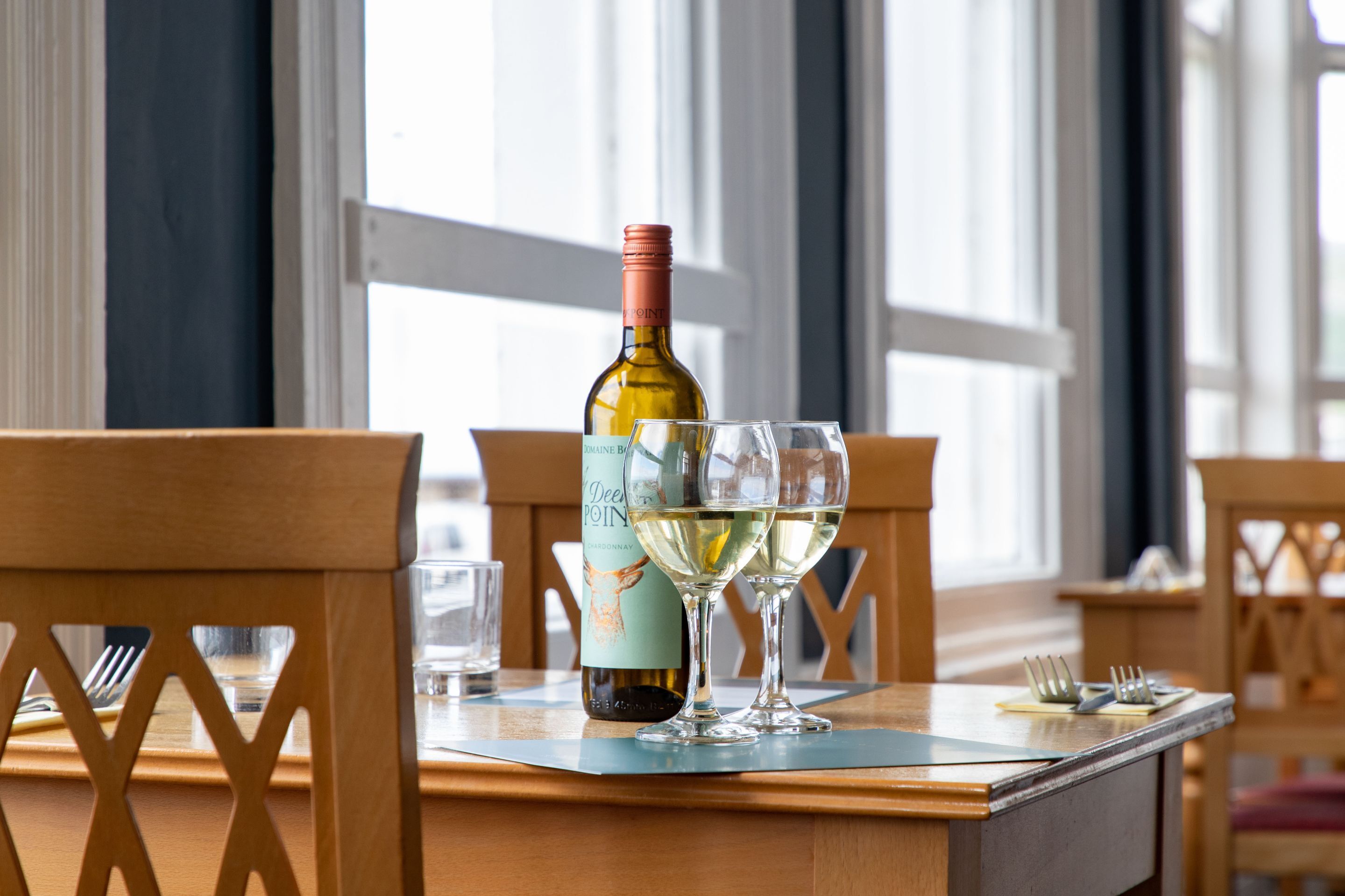 A table with a bottle of wine and two glasses on a wooden table in a room with large windows.