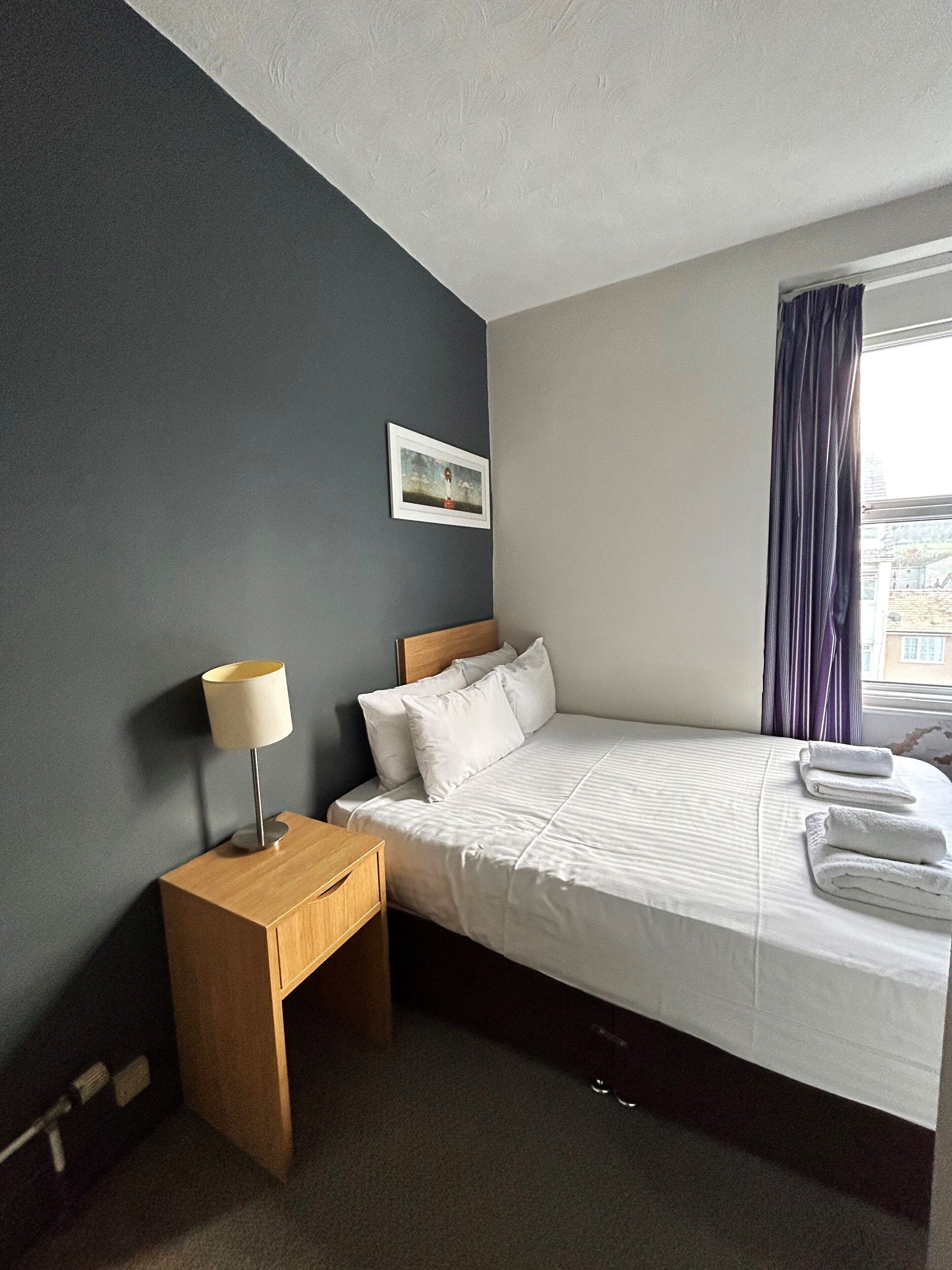 A hotel room with a bed, a nightstand, a lamp, a framed picture on the wall, and a window with curtains.