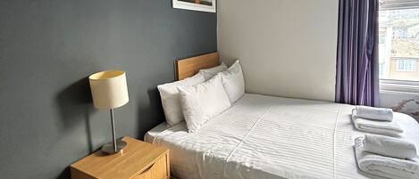 A hotel room with a bed, a nightstand, a lamp, a framed picture on the wall, and a window with curtains.