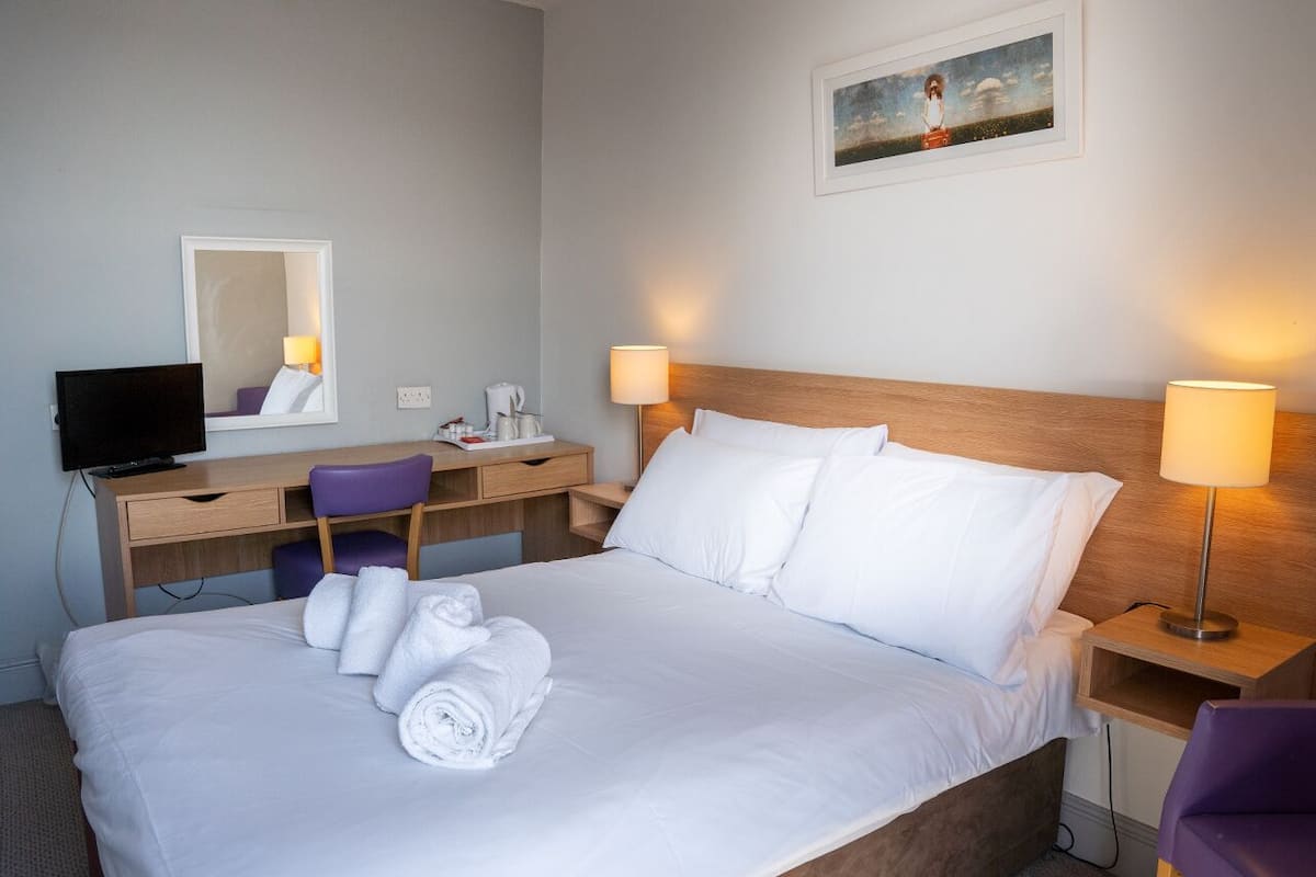 A neatly made bed with white linens and rolled towels, a wooden headboard, two bedside lamps, a desk with a chair, a television, and a painting on the wall.