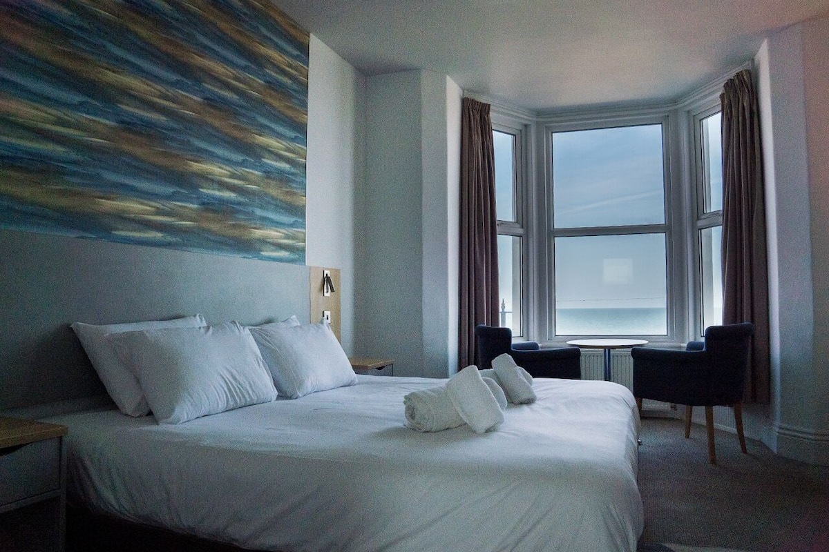A hotel room with a large bed, two chairs, a window with curtains, and a painting on the wall.
