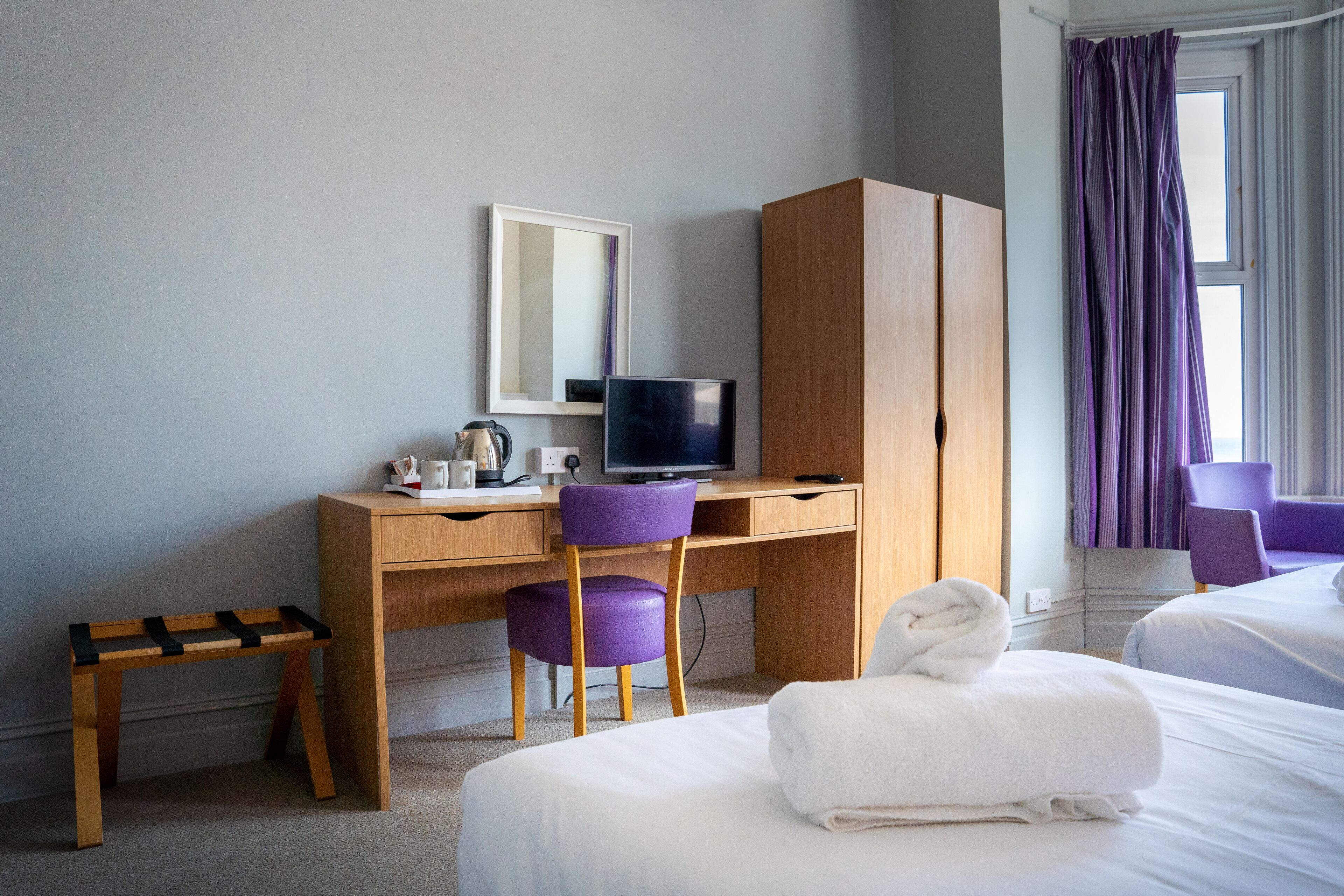 Ein Hotelzimmer mit einem Holztisch, einem violetten Sessel, einem Fernseher, einem kleinen Holzschemel und einem Bett mit einem weißen Handtuch.