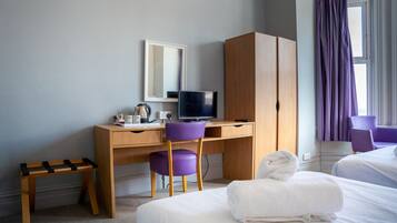 A hotel room with a wooden desk, a purple chair, a television, a small wooden stool, and a bed with a white towel.