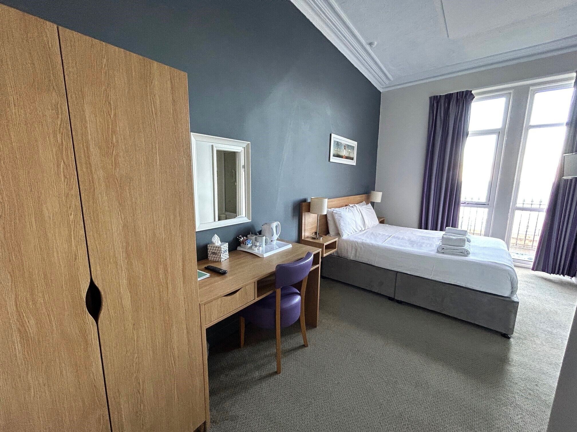 A hotel room with a bed, a desk, a chair, a wardrobe, a window, and a picture on the wall.
