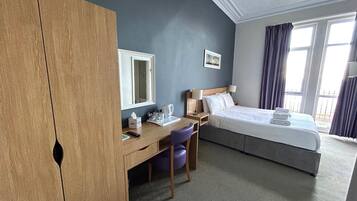 Quarto de hotel com uma cama, uma escrivaninha, uma cadeira, um guarda-roupa, uma janela e um quadro na parede.