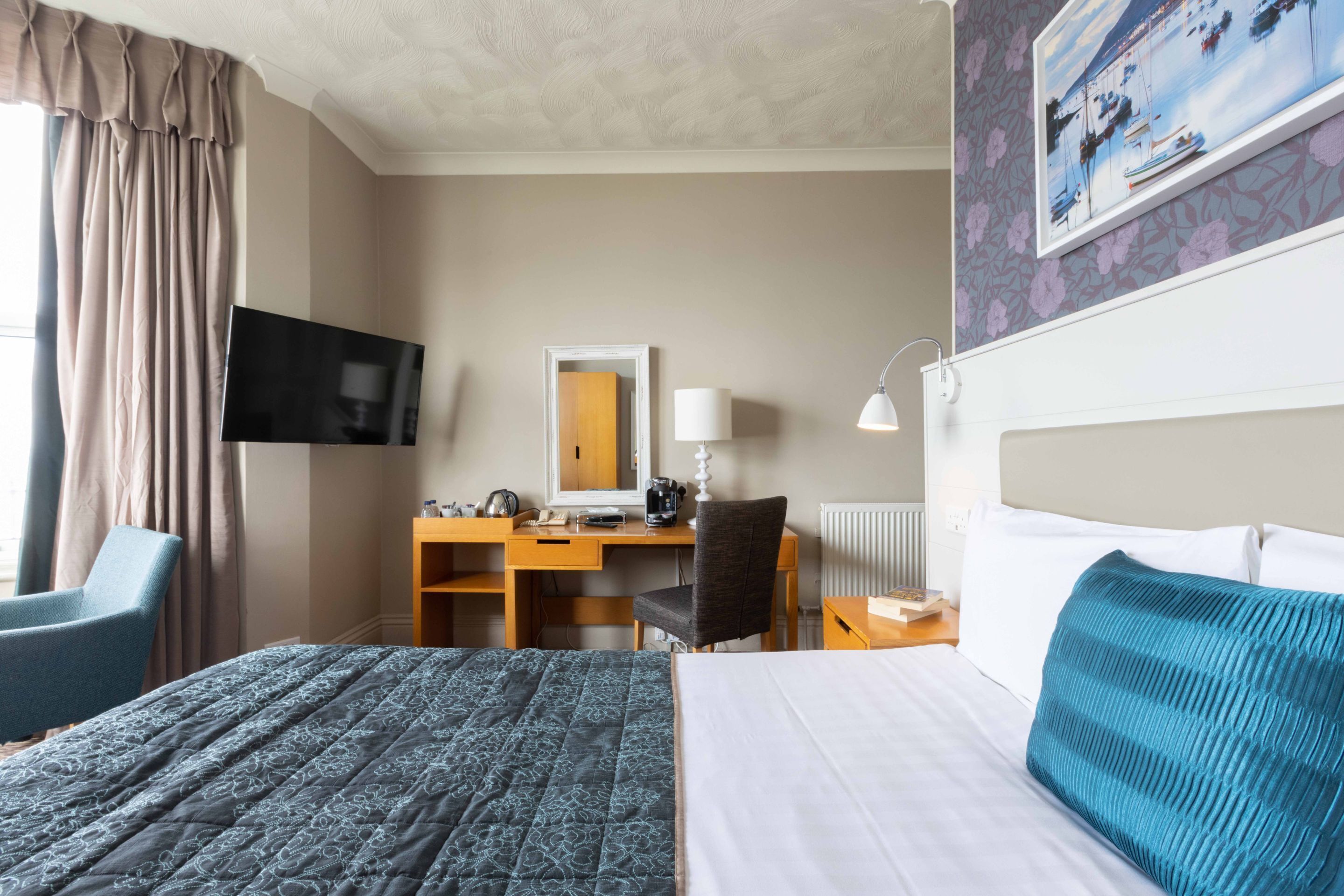 A hotel room with a bed, a desk with a chair, a TV, a lamp, and a painting on the wall.