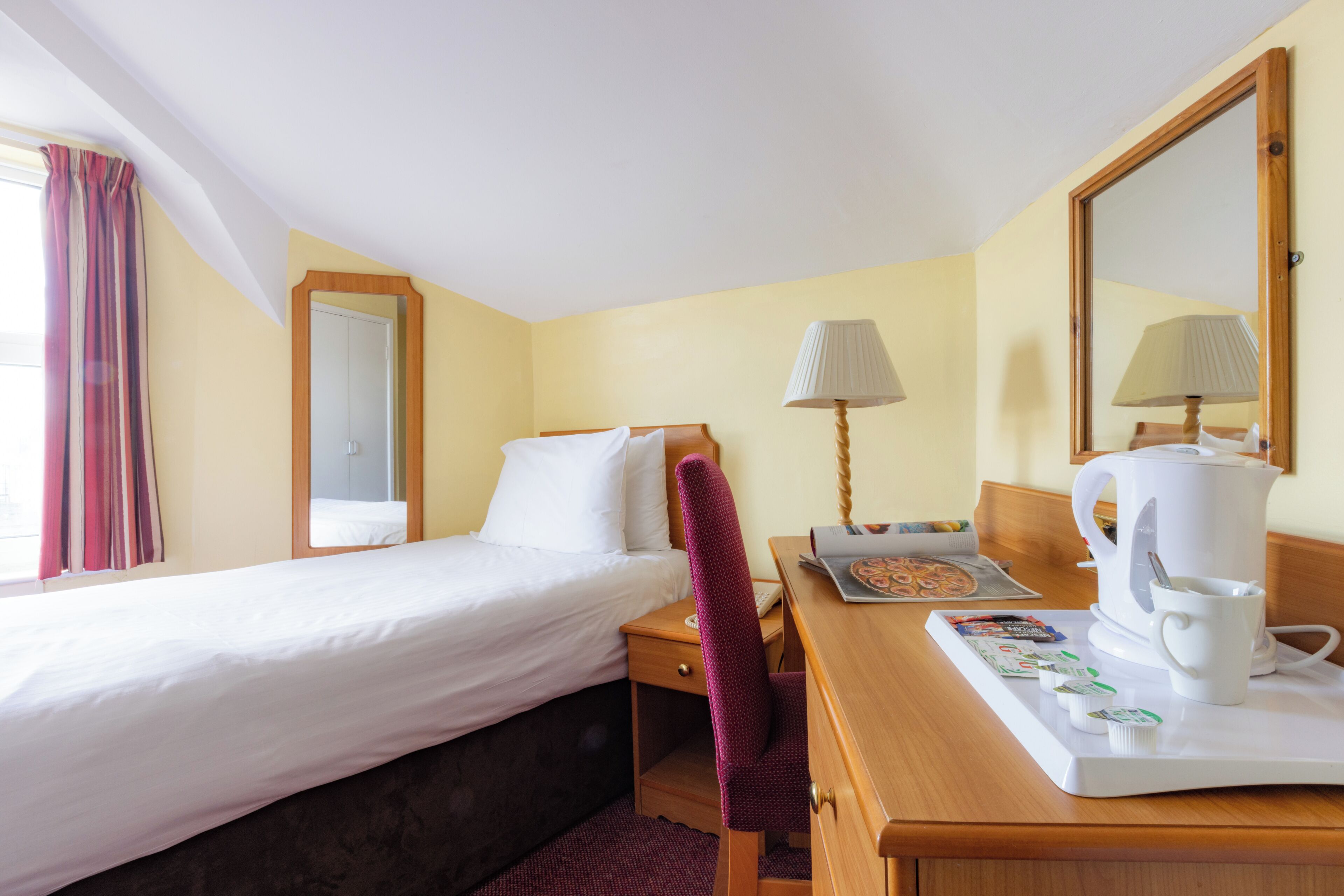 A hotel room with a bed, a desk with a coffee maker, a chair, a lamp, and a mirror.