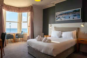 A hotel room with a large bed, two bedside tables, a desk with a chair, a window with curtains, and a painting on the wall. - Belvilla Llandudno Seafront Hotel (Llandudno)
