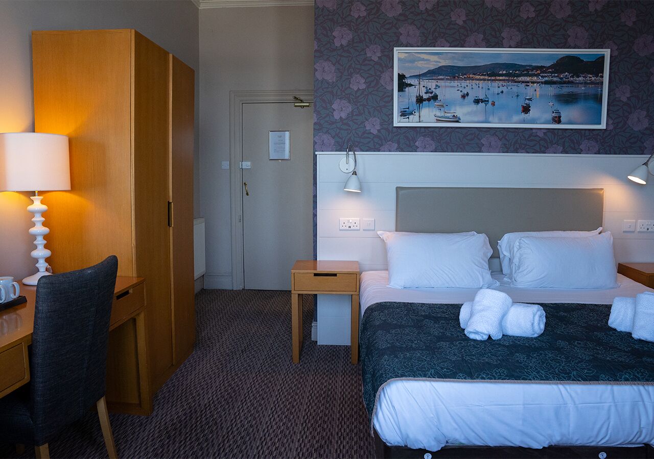 Quarto de hotel com cama, luminárias de cabeceira, escrivaninha com cadeira, mesinha e um quadro na parede.
