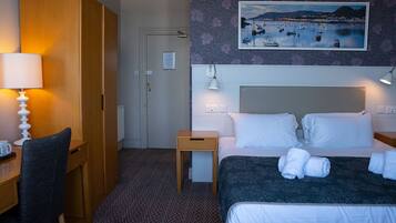 Quarto de hotel com cama, luminárias de cabeceira, escrivaninha com cadeira, mesinha e um quadro na parede.