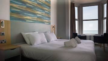 Habitación de hotel con una cama grande, vistas al mar a través de las ventanas, un cuadro en la pared y una pequeña mesa con una silla.