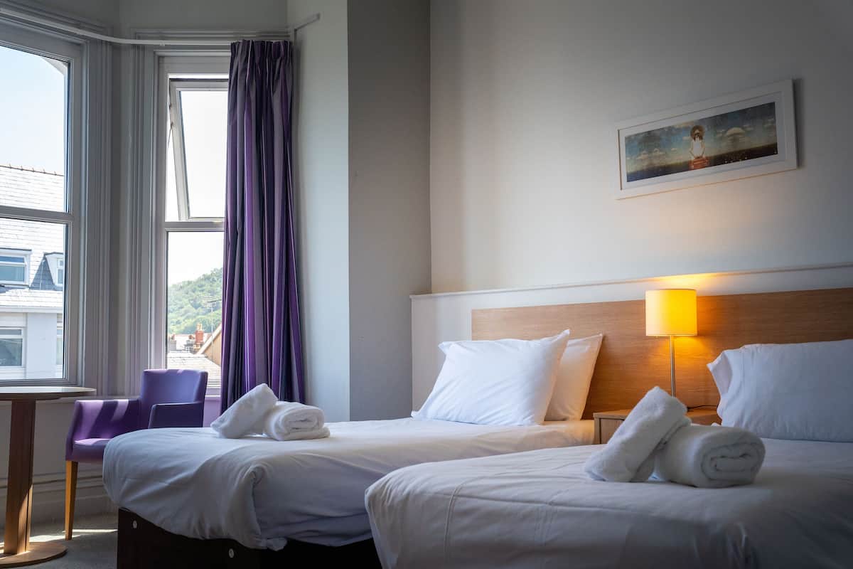 A hotel room with two beds, a window with curtains, a painting on the wall, and a lamp.