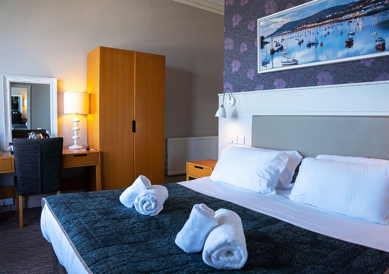 A hotel room with a bed, a desk, a chair, a wardrobe, and a painting of a harbor scene.