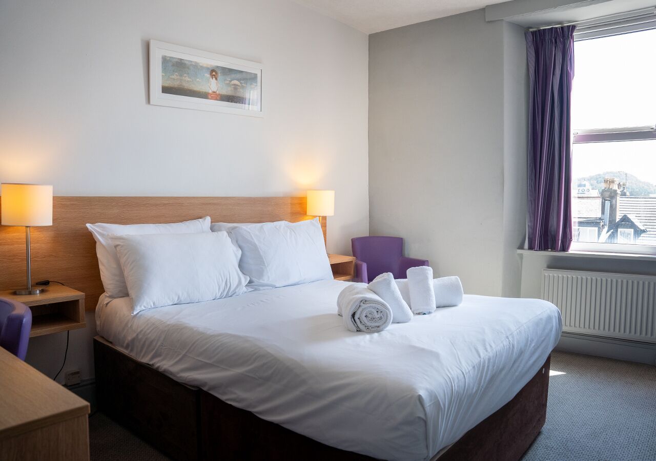 Una camera d'albergo con un letto grande, due abat-jour, una sedia e una finestra con tende.